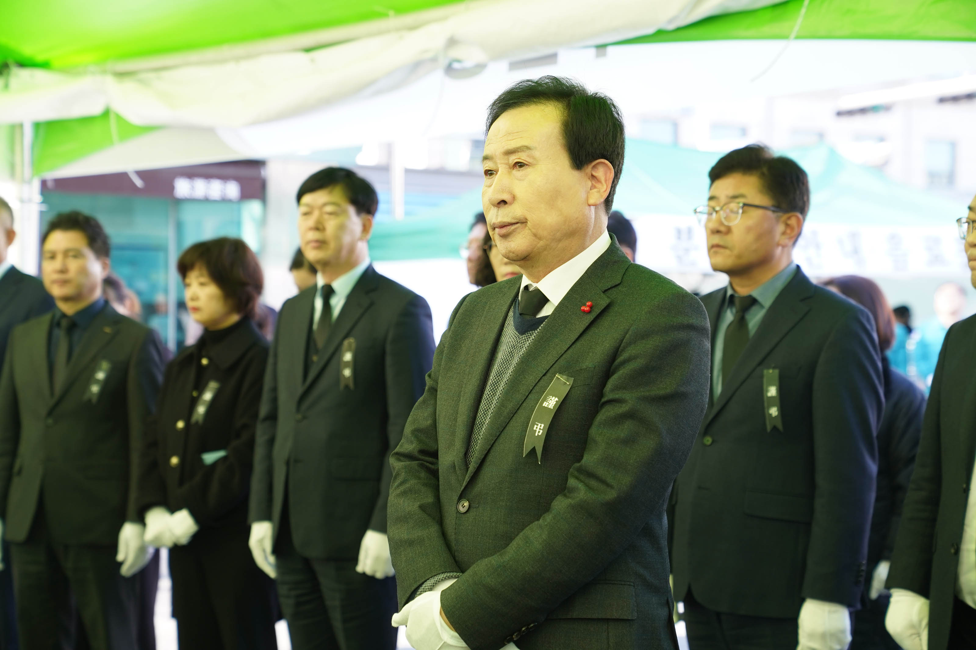 가슴에 검정 리본을 단 박홍률 목포 시장이 조문 행사에서 엄숙한 표정으로 서 있다. 주변에는 흰 장갑을 낀 다른 조문객들도 함께 서 있으며, 모두 슬픔을 간직한 채 고개를 숙이거나 앞을 바라보고 있다. 배경에는 초록색 천막과 분향소 안내 문구가 적힌 천막이 보인다. 전체적으로 차분하고 엄숙한 분위기다.
