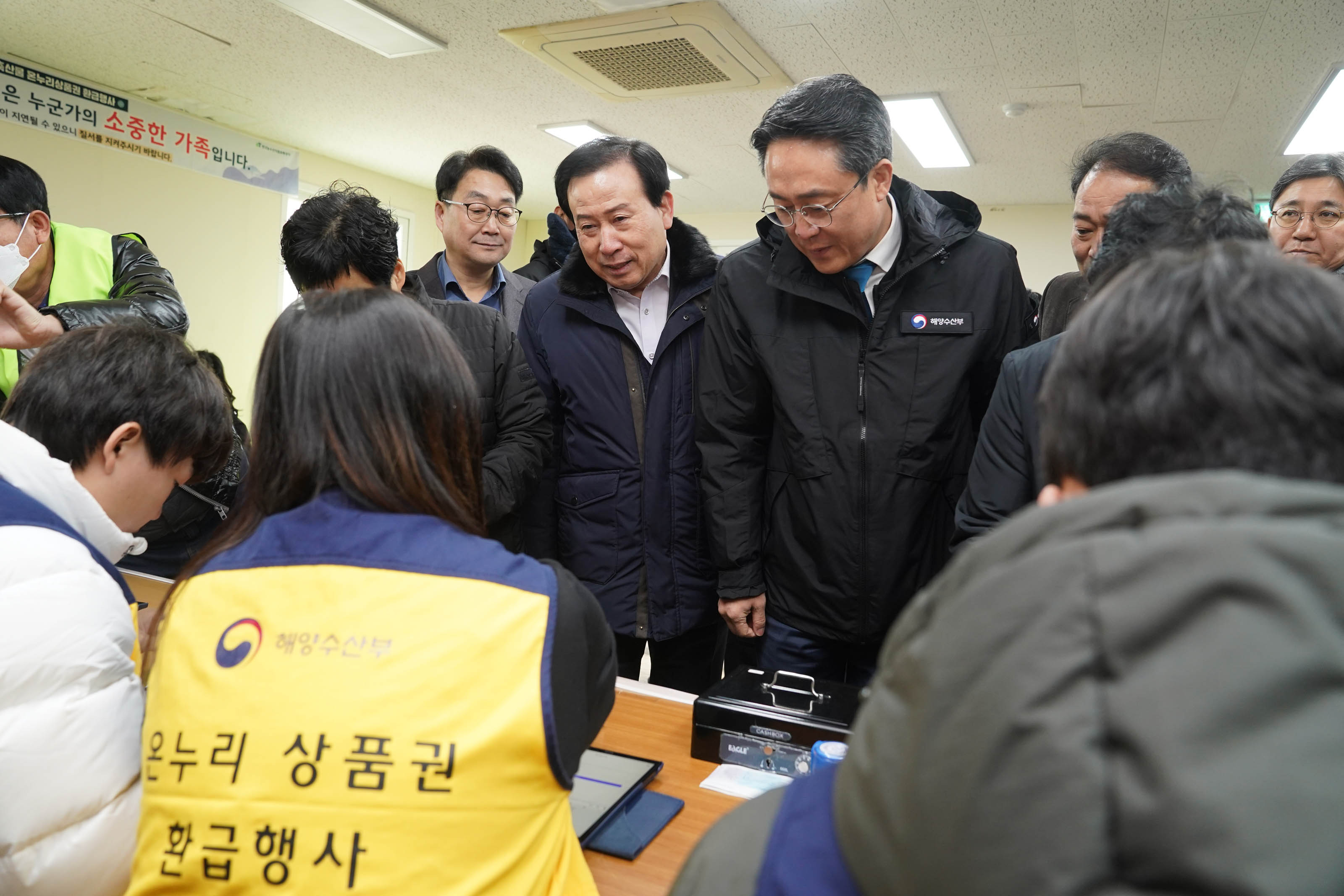 실내 행사장에서 박홍률 목포시장과 강도형 해수부장관 그리고 관계자들이 온누리상품권 환급 부스를 방문하고 있다. 앞쪽에는 노란 조끼를 입은 직원이 앉아 있으며, 조끼 등판에는 '해양수산부 온누리 상품권 환급행사' 문구가 적혀 있다.
