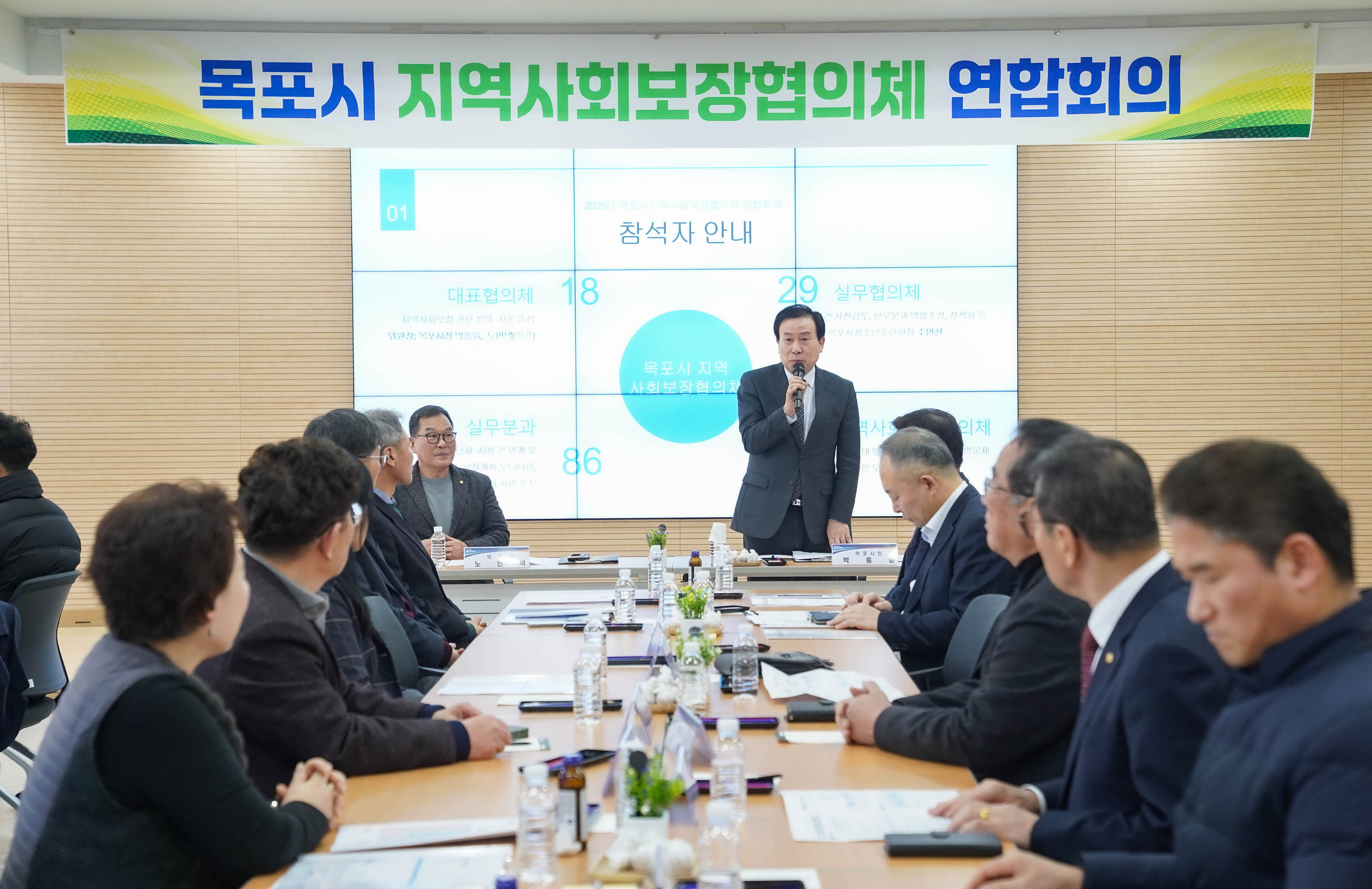 목포시 지역사회보장협의체 연합회의 행사장에서 양복을 입은 박홍률 목포시장이 무선 마이크를 들고 발언 중인 모습. 참석자들은 긴 테이블 양쪽에 나뉘어 앉아 발언을 경청하고 있음. 뒤 전광판 화면에는 '참석자 안내', '대표협의체 18', '실무협의체 29', '실무분과 86', '동 지역사회보장협의체 390' 등의 항목과 함께 '목포시 지역사회보장협의체'라는 파란 원이 큼직하게 표시되어 있음. 상단 현수막에는 '목포시 지역사회보장협의체 연합회의' 문구가 강조되어 있음.