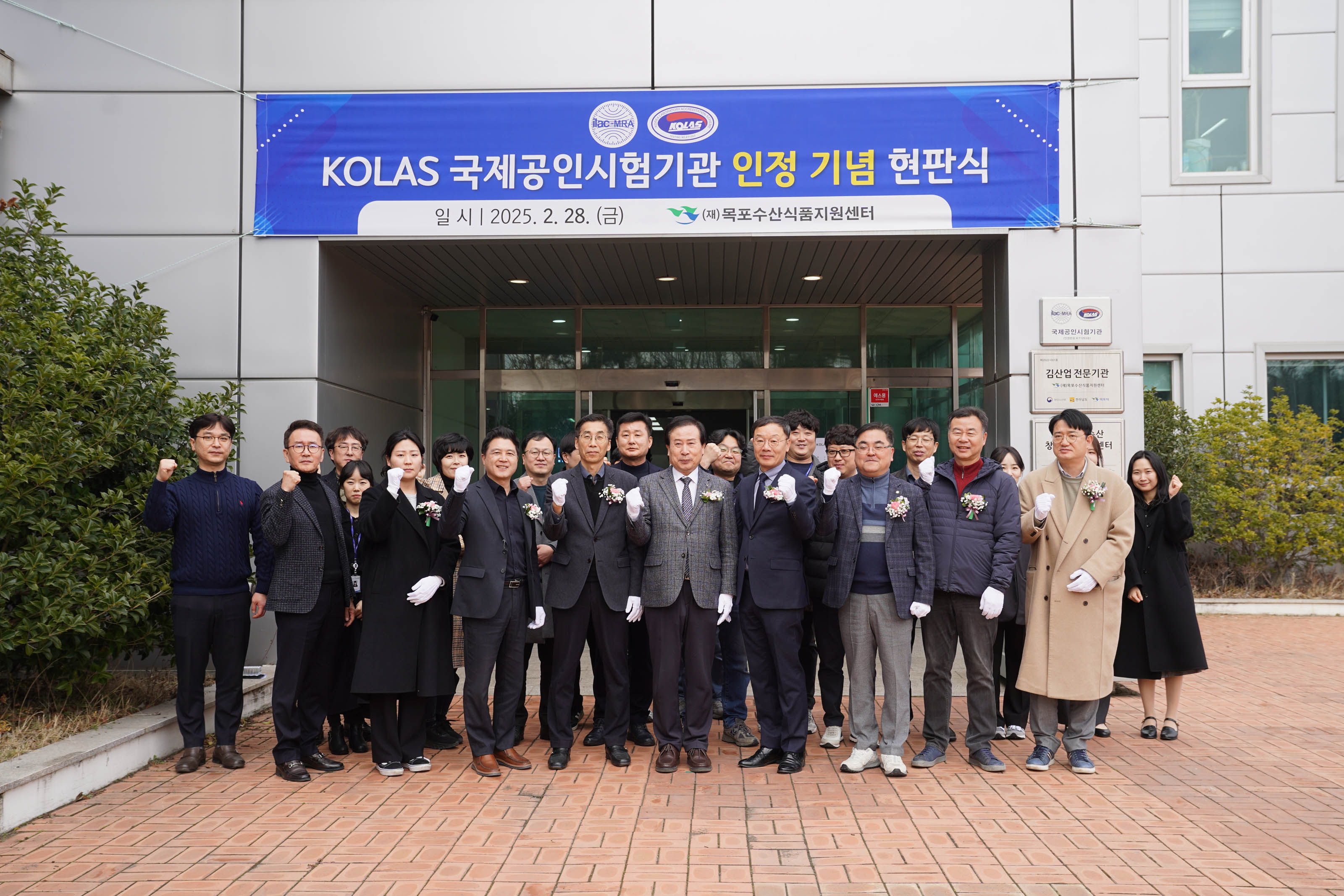 박홍률 목포시장을 비롯한 많은 인원이 단체로 파이팅 포즈를 하며 기념촬영을 하고 있음. 모두 검은 옷 또는 정장을 입고 흰 장갑 착용. 건물 출입문 위에 파란색 현수막이 걸려 있고 텍스트는 다음과 같음:
'KOLAS 국제공인시험기관 인정 기념 현판식
일 시 | 2025. 2. 28. (금)
(재)목포수산식품지원센터'