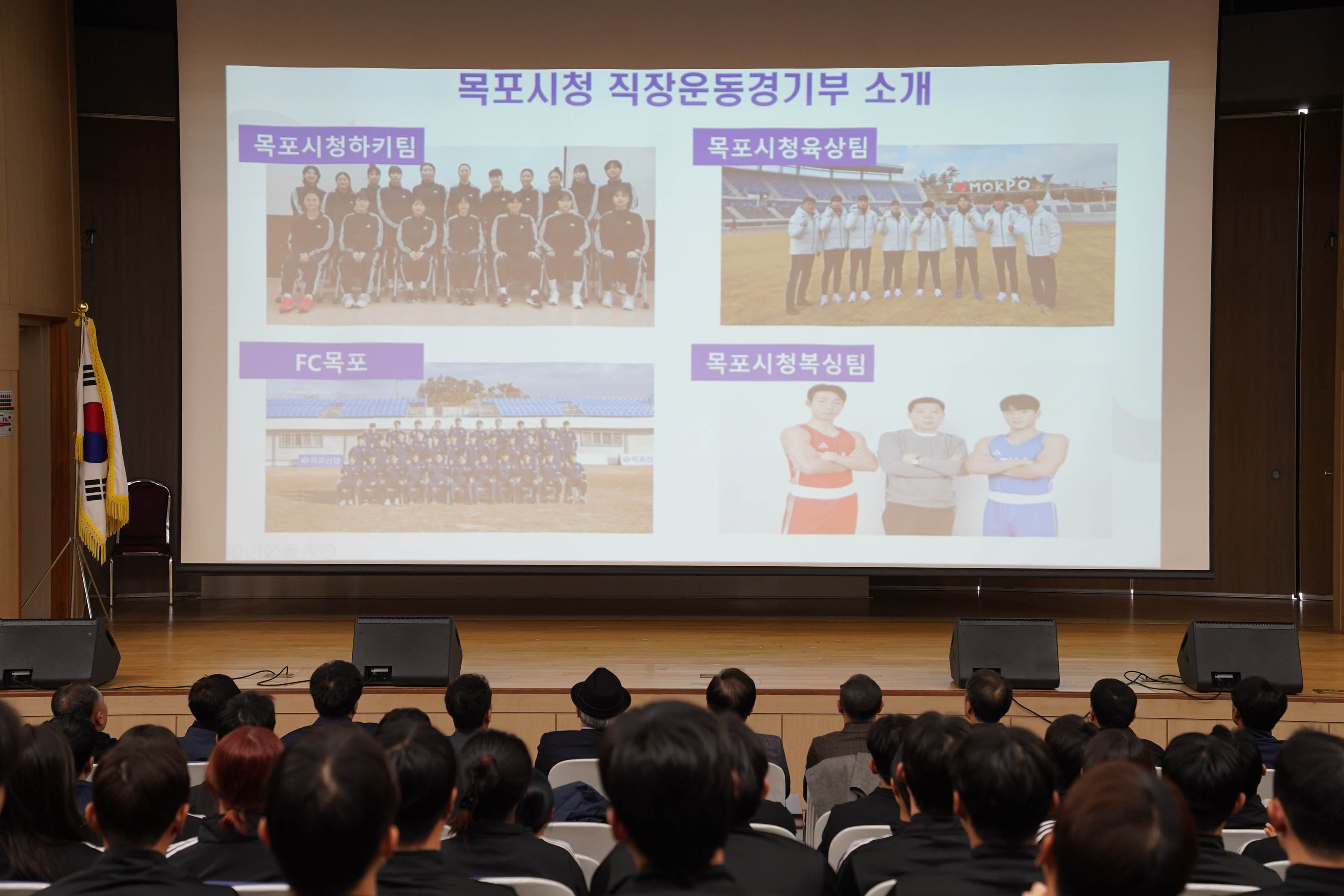 목포시청 직장운동경기부를 소개하는 프레젠테이션 슬라이드. 화면에는 네 개의 운동팀 사진이 있고, 각각 목포시청하키팀, 목포시청육상팀, FC목포, 목포시청복싱팀이라는 텍스트가 있다. 객석에는 많은 사람들이 슬라이드를 보고 있음. 무대 옆에는 태극기가 놓여 있고, 목포시 로고도 보임.