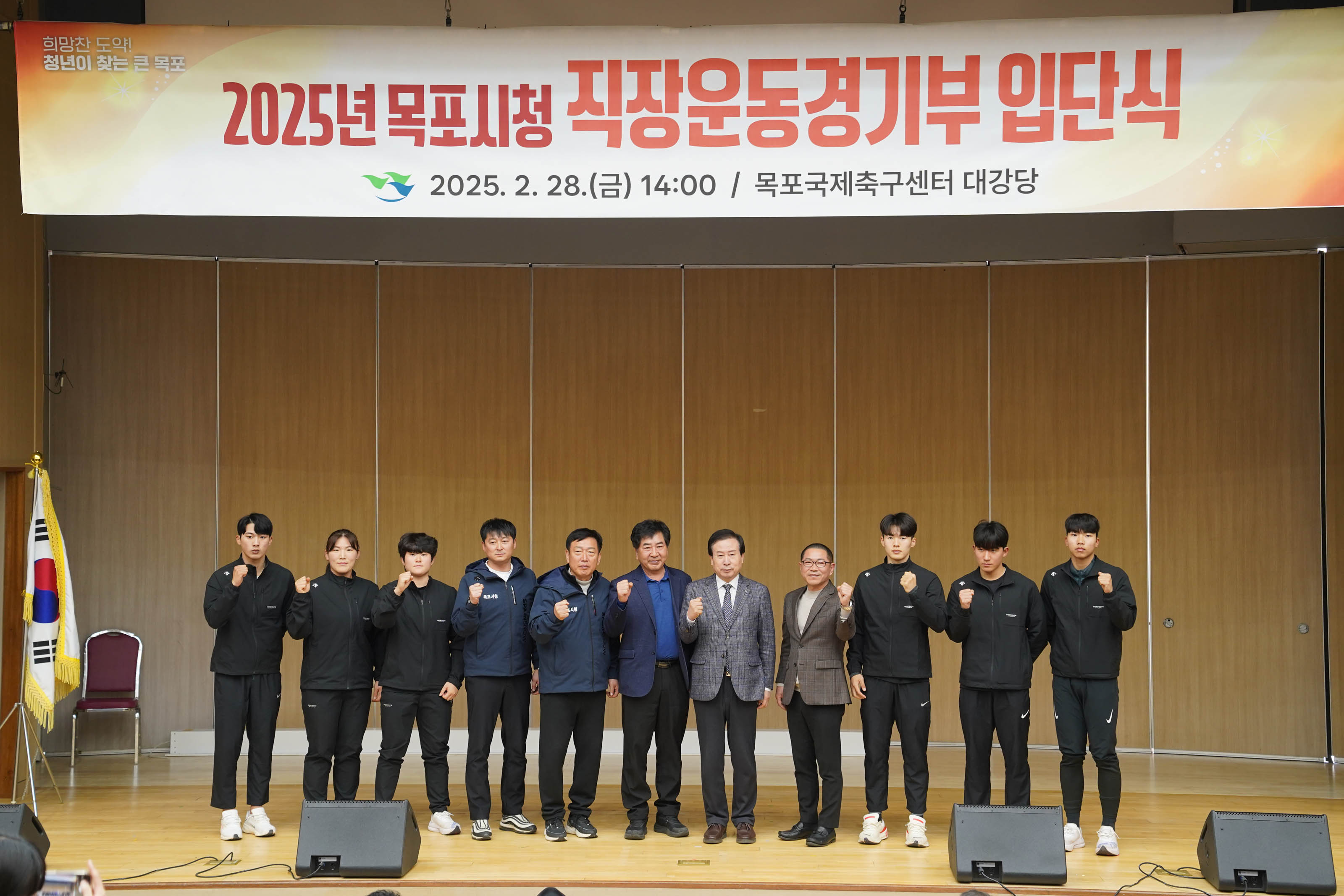 박홍률 목포시장을 비롯한 여러 명의 참가자가 무대 중앙에서 파이팅 포즈를 취하며 단체 사진을 찍고 있음. 위에는 '2025년 목포시청 직장운동경기부 입단식 2025. 2. 28.(금) 14:00 / 목포국제축구센터 대강당' 현수막이 걸려 있으며, FC목포 및 기타 종목 관계자로 보이는 인물들이 포함됨.