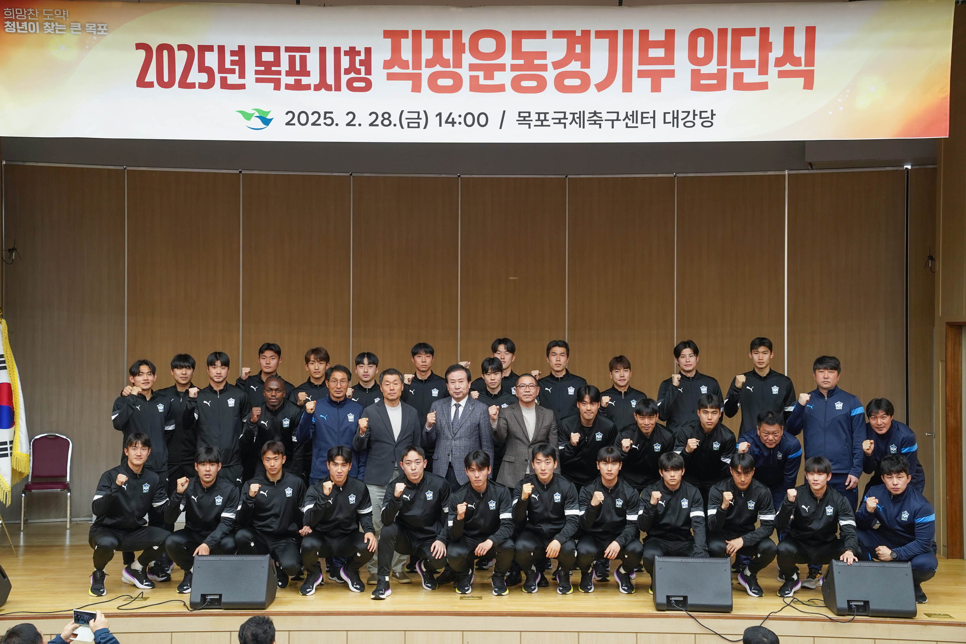 FC목포 소속으로 보이는 유니폼을 입은 많은 운동선수들과 박홍률 목포시장을 비롯한 관계자들이 무대 위에서 단체 사진을 찍고 있으며, 모두 파이팅 포즈를 취하고 있음. 무대 위에는 ‘2025년 목포시청 직장운동경기부 입단식 2025. 2. 28.(금) 14:00 / 목포국제축구센터 대강당’ 이라는 현수막이 걸려 있고, 태극기와 목포시 로고도 보인다.