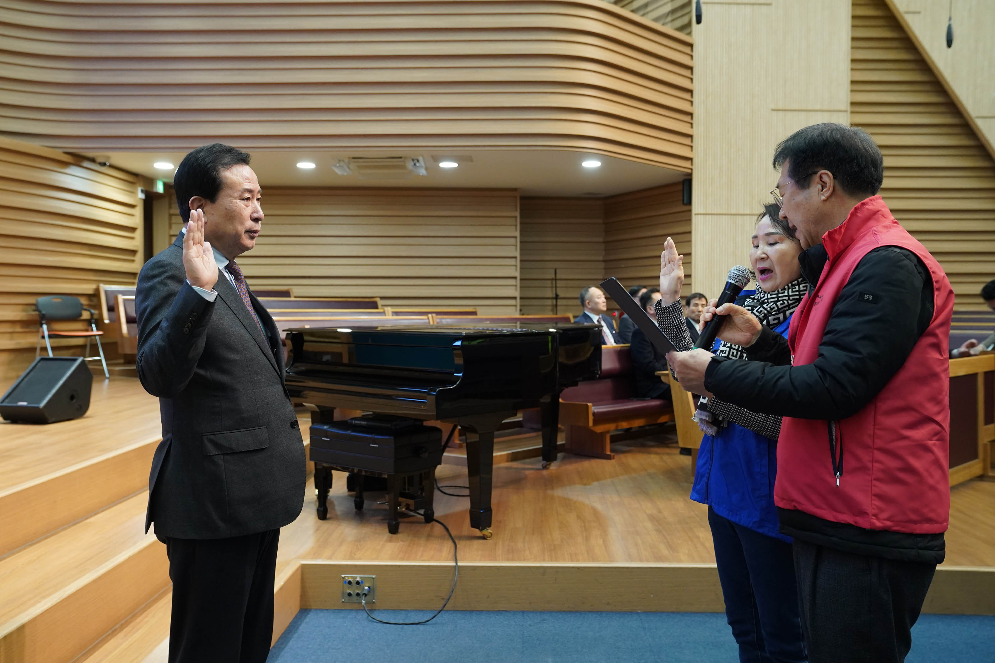 실내 무대 앞에서 남녀 두 명이 마이크를 들고 선서를 하고 있고, 박홍률 목포시장은 오른손을 들고 마주보면서 선서를 받고 있다. 무대 뒤쪽에는 검정색 그랜드 피아노와 예배당 의자들이 배치되어 있으며, 주변에서는 정장을 입은 관계자들이 앉아 지켜보고 있다.