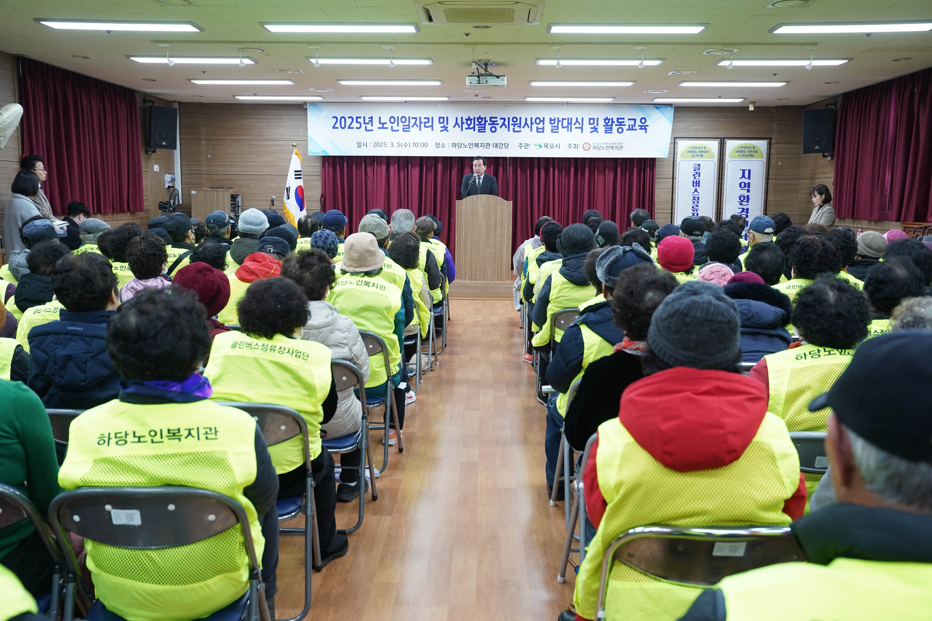 하당노인복지관 강당 전경. 중앙 무대에 한 관계자가 연단에서 마이크로 발언하고 있고, 참석자들이 등 돌리고 앉아 강연을 경청하고 있다. 대부분 형광 노란색 조끼를 착용한 노년층이며, 조끼 뒤에는 '하당노인복지관' 등의 문구가 새겨져 있다. 무대 위 현수막에는 '2025년 노인일자리 및 사회활동지원사업 발대식 및 활동교육 일시: 2025. 3. 5(수) 10:00 장소: 하당노인복지관 대강당 주관: 목포시 주최: 하당노인복지관'이 명시되어 있으며, 오른쪽에는 '클린버스정류장사업단', '지역환경개선'이라는 세로형 안내판이 보인다.