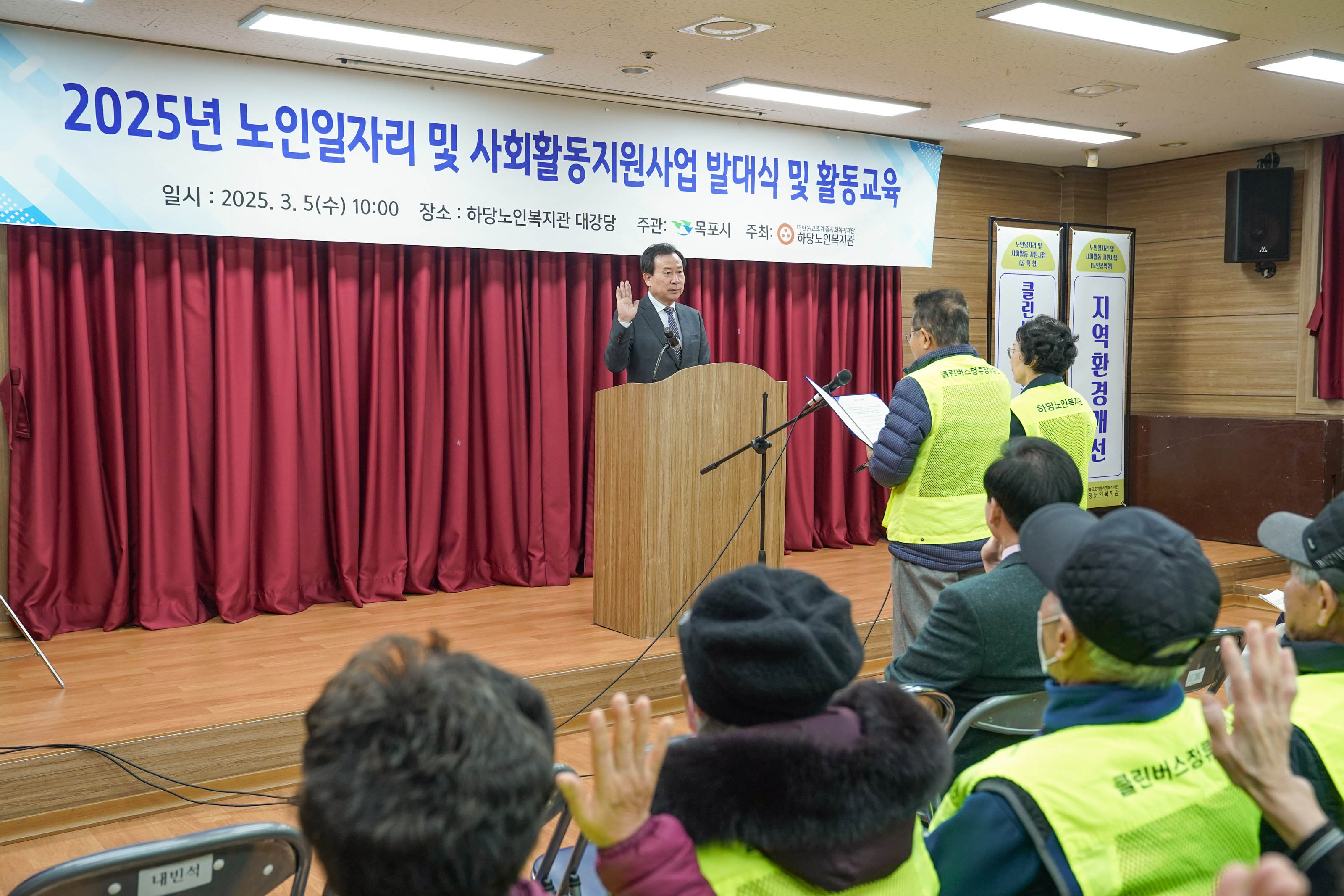 하당노인복지관 대강당 무대에서 박홍률 목포시장이 나무 연단에 서서 오른손을 들고 선서를 하고 있다. 연단 앞에는 마이크 두 개가 설치되어 있으며, 앞쪽에는 형광 노란 조끼를 입은 두 명의 참여자가 종이를 들고 선서를 진행하고 있다. 무대 상단에는 흰색 현수막이 걸려 있으며, 그 위에는 '2025년 노인일자리 및 사회활동지원사업 발대식 및 활동교육 일시 : 2025. 3. 5(수) 10:00 장소 : 하당노인복지관 대강당 주관: 목포시 주최: 하당노인복지관'이라는 문구가 적혀 있다. 우측에는 '클린버스', '지역환경개선'이라는 세로형 안내 배너가 보인다.