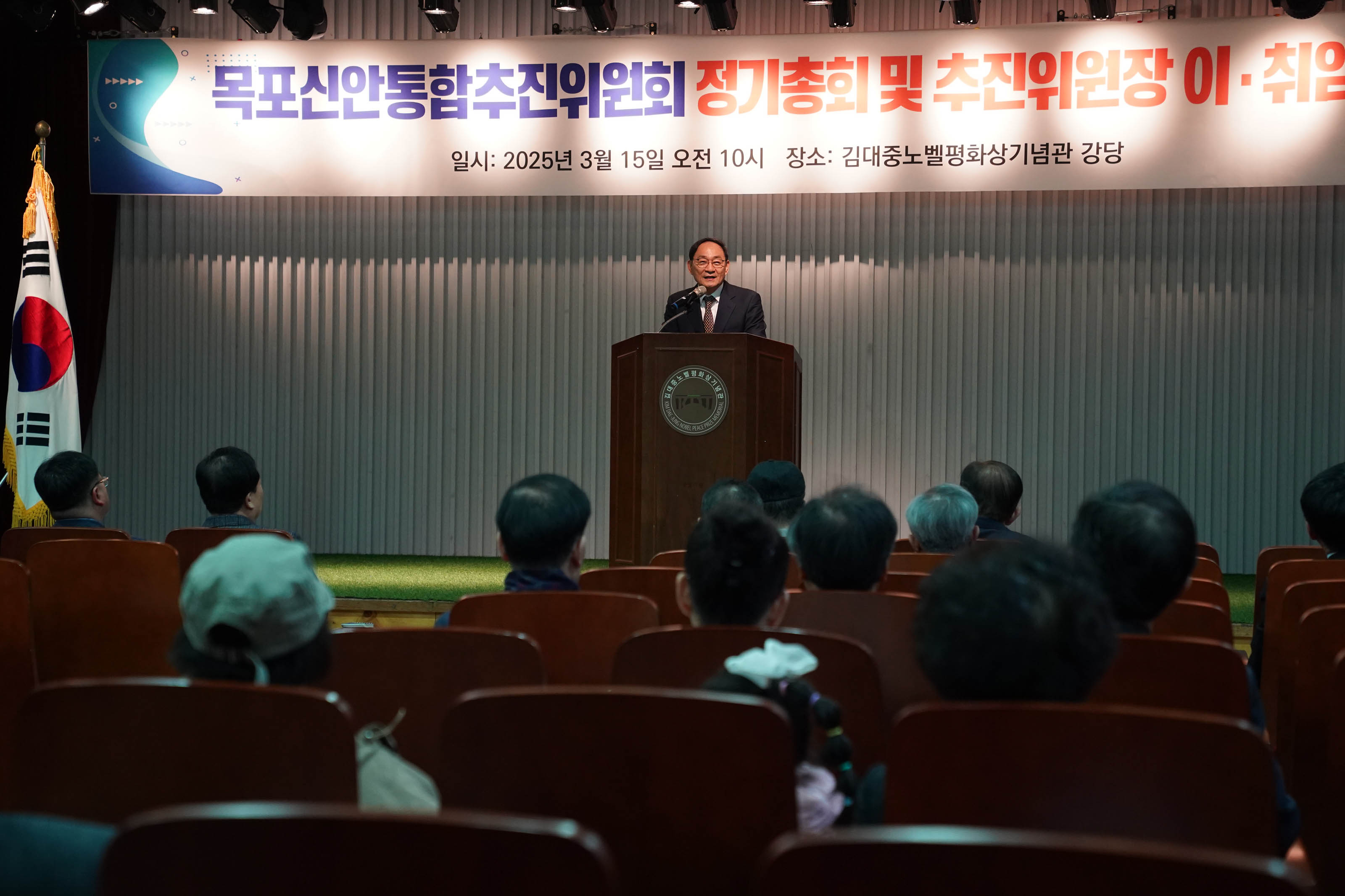 목포신안통합추진위원회 정기총회 및 추진위원장 이·취임식에서 관계자가 연단에서 발언하고 있다. 배경 배너에는 '목포신안통합추진위원회 정기총회 및 추진위원장 이·취임식'이라는 문구와 함께 '일시: 2025년 3월 15일 오전 10시', '장소: 김대중노벨평화상기념관 강당'이 적혀 있다. 청중들이 연단을 향해 앉아 있다.