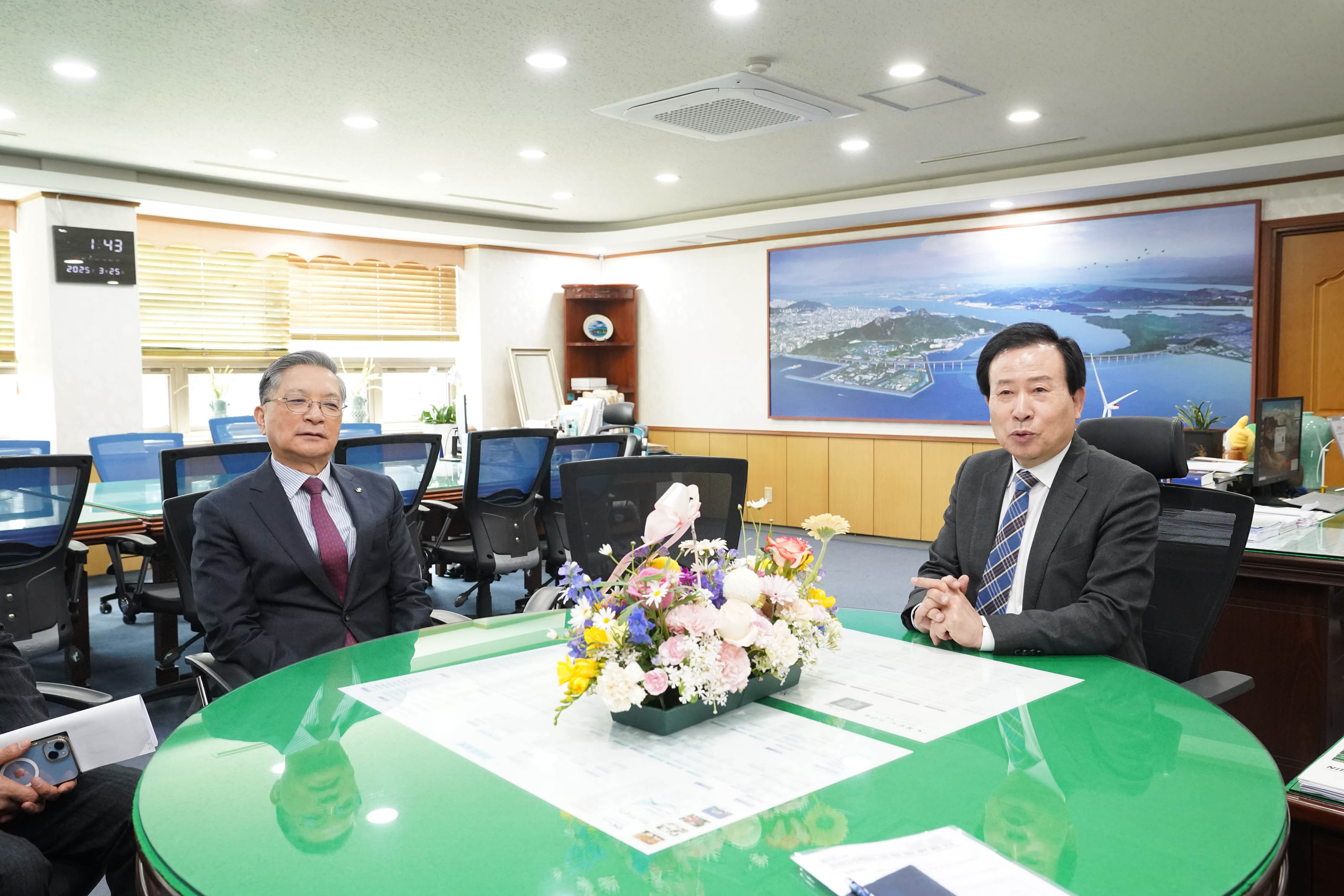 사무실에서 원형 회의 테이블을 사이에 두고 양복을 입은 박홍률 목포시장과 이한준 한국토지주택공사 사장이 대화하고 있다. 테이블 위에는 꽃바구니가 놓여 있고, 벽 시계에는 '1:43 / 2025. 3. 25'라고 표시되어 있다. 배경에는 항공 촬영된 도시 전경 사진이 걸려 있다.