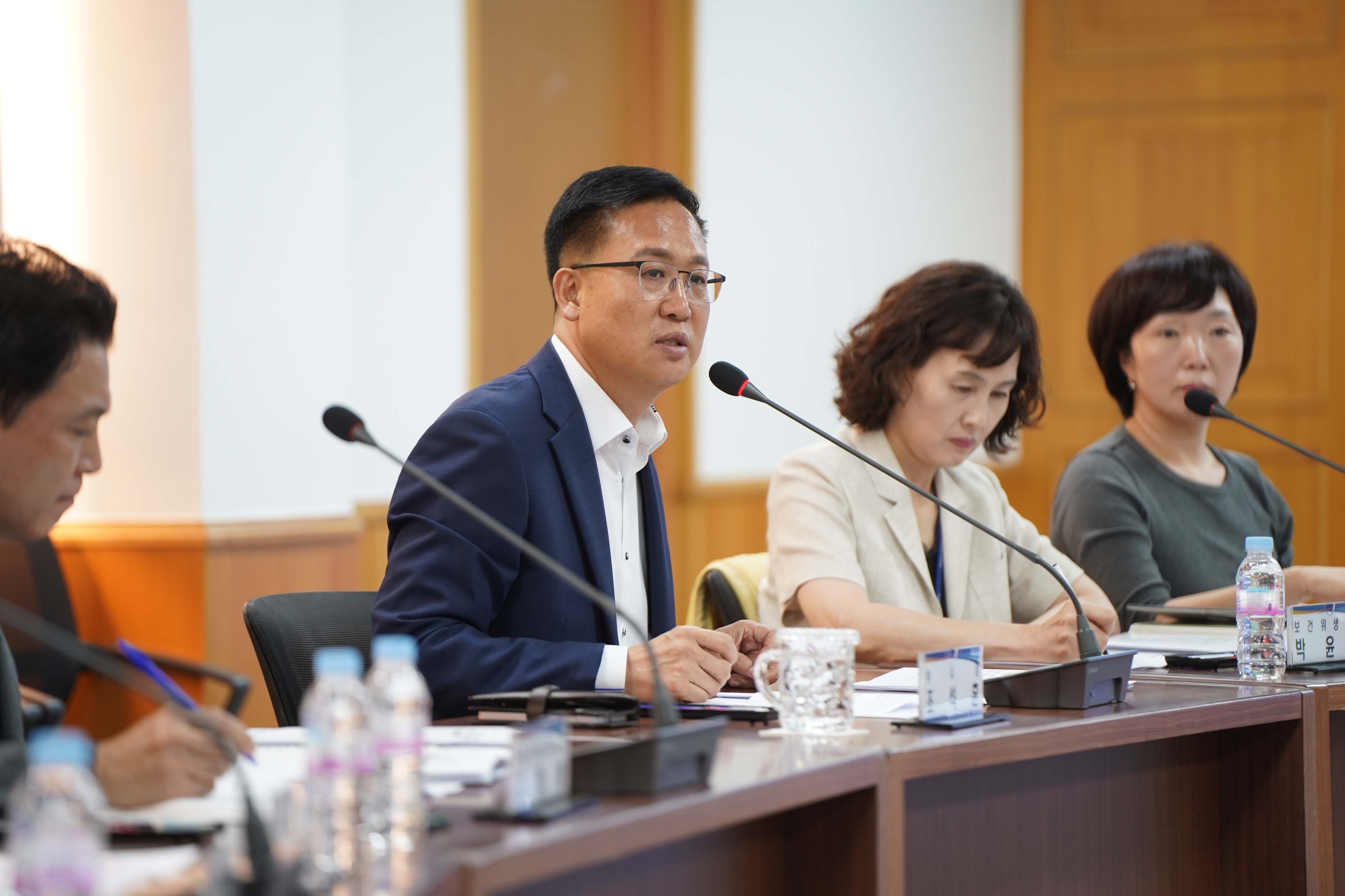 회의 테이블 중앙에 앉아 마이크 앞에서 발언하고 있는 남성의 근접 사진. 남성 오른쪽으로 두 명의 여성이 앉아 있으며, 테이블 위에는 물병과 컵이 놓여 있음.
