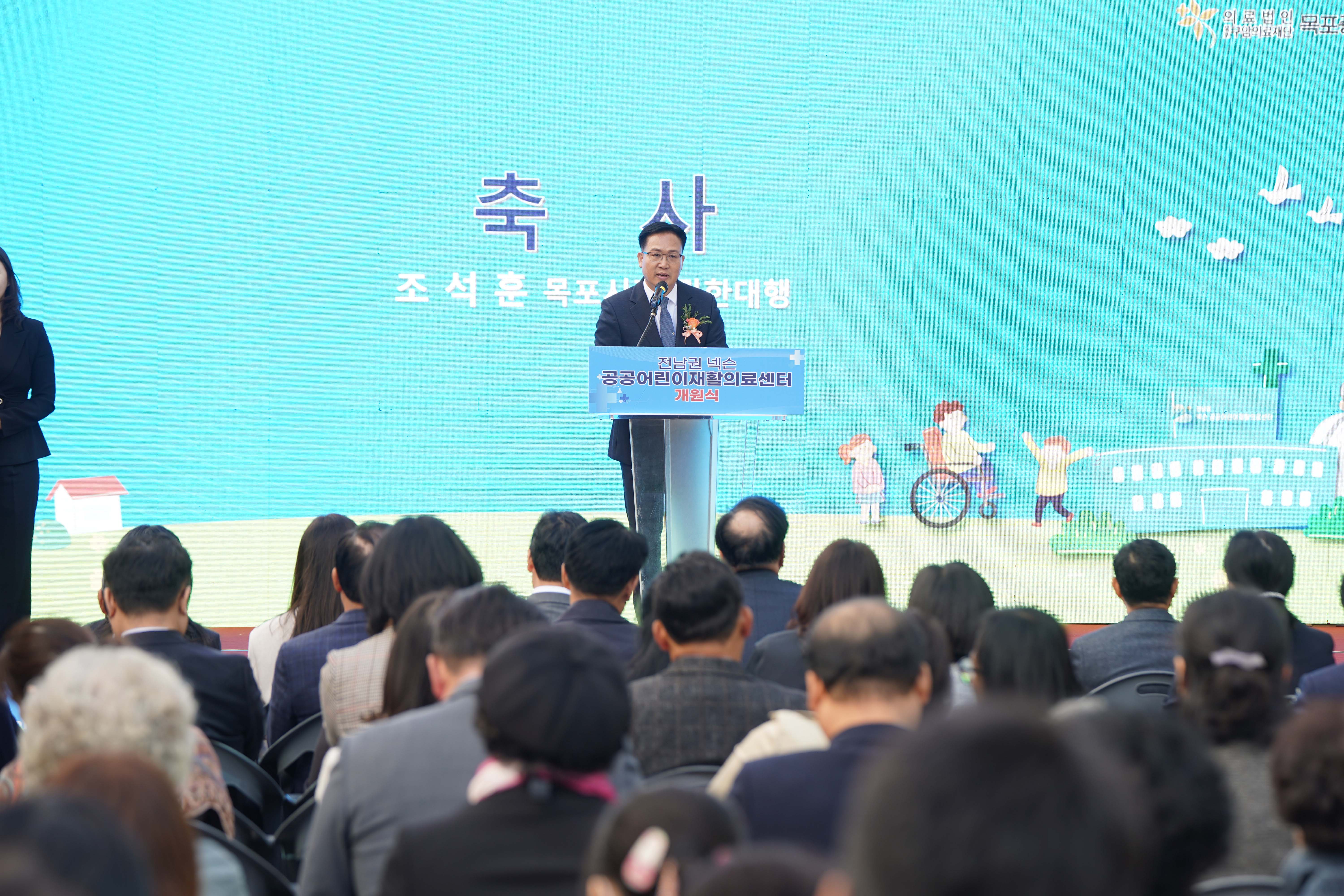 '전남권 넥슨 공공어린이재활의료센터 개원식' 무대에서 조석훈 목포시장 권한대행이 축사를 하는 모습. 배경 스크린에 '축사 조석훈 목포시장 권한대행' 텍스트가 보임.