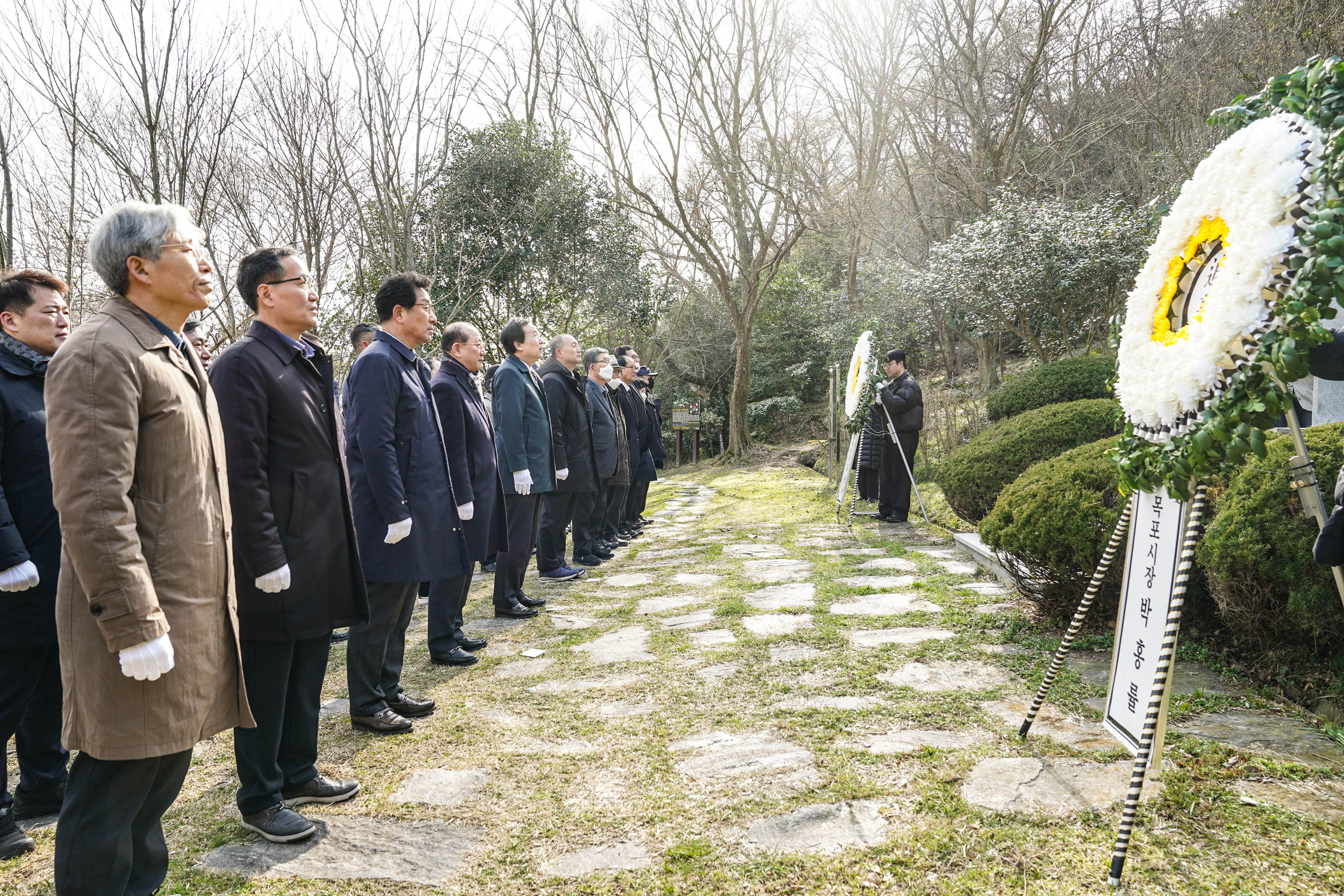 유달산3.1운동기념탑 앞에서 박홍률 시장을 비롯한 대략 20명 정도의 사람들이 흰색 장값을 끼고 가로 두줄로 나란히 서있다. 기념찹 앞에 동그란 모양의 하얀색 조화 두개가 보인다. 목포시장 박홍률이라고 적힌 조화가 가까이 보인다.