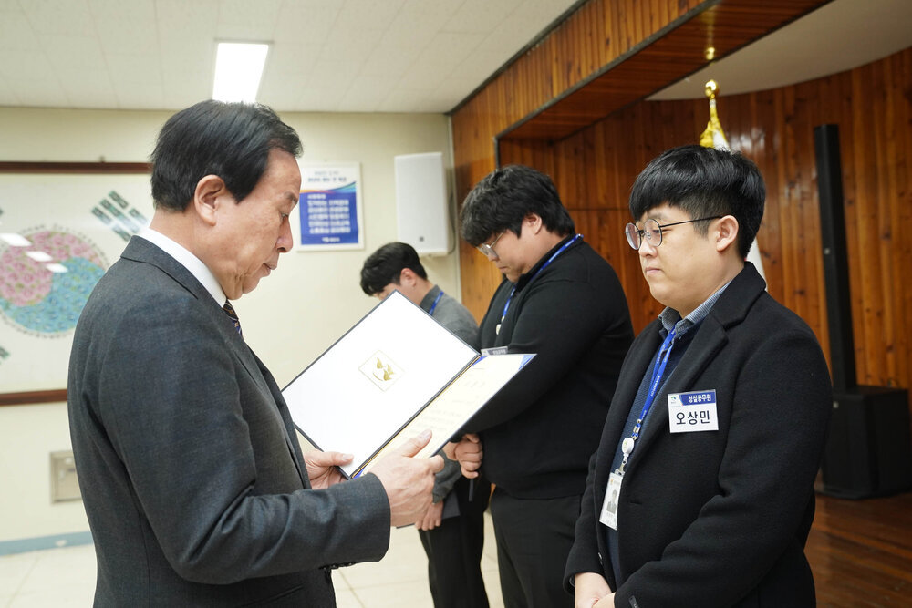2월 정례조회에서 정장입은 남성이 검정 자켓을 입은 남성에게 상장을 건내고 있다.