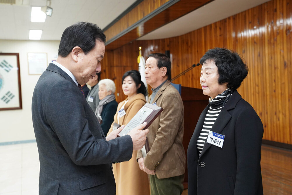 3월 정례조회에서 정장입은 남성이 검은색 자켓을 입은 여성에게 패를 건내고 있다.