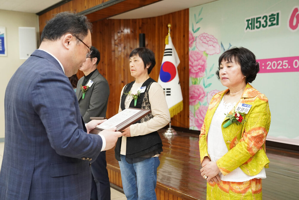 정장을 입고 안경을 낀 남성이 노란색 자켓에 오렌지 무늬가 있는 투피스를 입은 장영미에게 패를 수여하고 있다