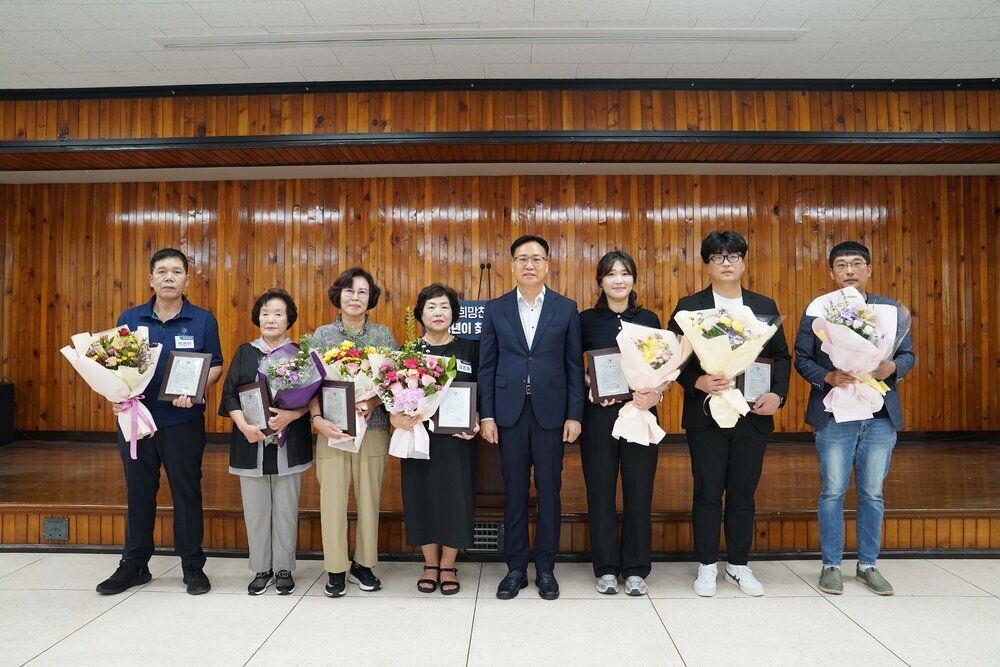 표창패 수여식에서 정장과 단정한 복장의 수상자들이 꽃다발과 패를 들고 단체 기념 촬영을 하고 있는 모습이다.