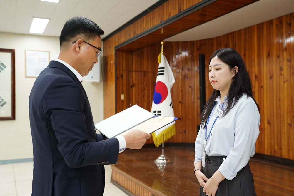 정장 차림의 관계자가 하늘색 블라우스를 입은 여성 수상자에게 상장을 전달하며 읽고 있는 모습이다.