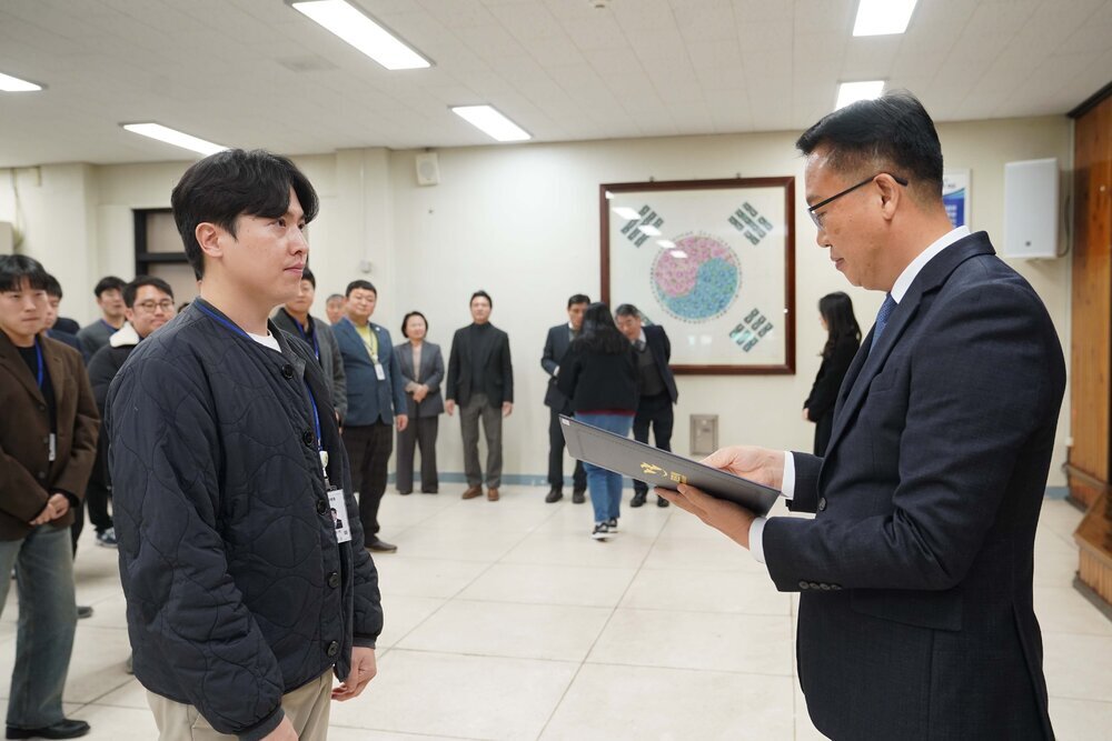 2026년 상반기 4급이하 정기인사로 정장을 입은 남성이 실내 행사장에서 한 남성에게 상장을 전달하며 내용을 읽고 있고, 남성은 검은색 퀼팅 자켓을 입고 사원증을 착용한 채 두 손을 내린 상태로 서 있으며 뒤쪽으로 참석자들이 줄지어 서서 행사를 지켜보고 있다.