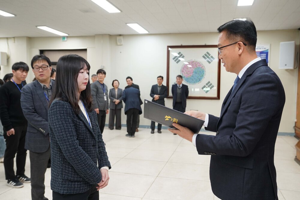 2026년 상반기 4급이하 정기인사로 정장을 입은 남성이 실내 행사장에서 한 여성에게 상장을 전달하며 내용을 읽고 있고, 여성은 남색 트위드 자켓을 입고 사원증을 착용한 채 두 손을 모은 상태로 서 있으며 뒤쪽으로 참석자들이 줄지어 서서 행사를 지켜보고 있다.