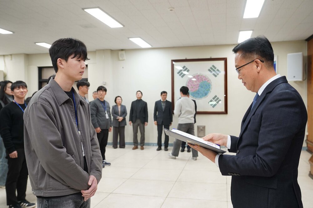 2026년 상반기 4급이하 정기인사로 정장을 입은 남성이 실내 행사장에서 한 남성에게 상장을 전달하며 내용을 읽고 있고, 남성은 회색 집업 점퍼를 입고 사원증을 착용한 채 두 손을 모은 상태로 서 있으며 뒤쪽으로 참석자들이 줄지어 서서 행사를 지켜보고 있다.