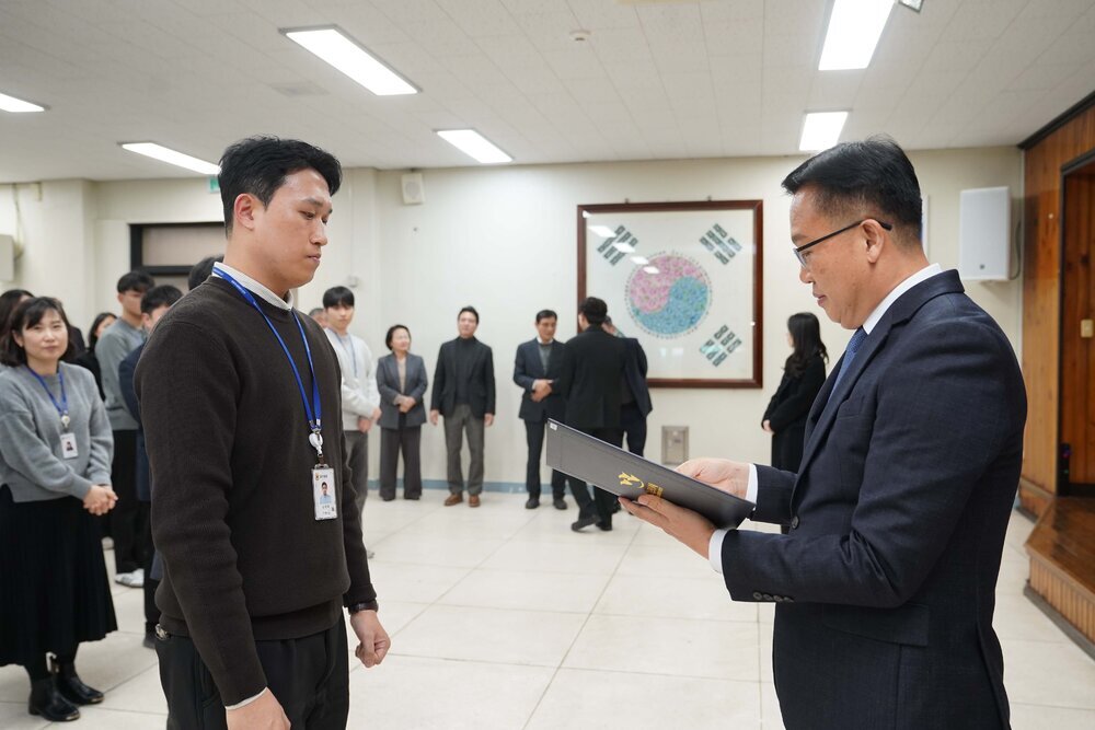 2026년 상반기 4급이하 정기인사로 정장을 입은 남성이 실내 행사장에서 한 남성에게 상장을 전달하며 내용을 읽고 있고, 남성은 갈색 니트를 입고 사원증을 착용한 채 두 손을 내린 상태로 서 있으며 뒤쪽으로 참석자들이 줄지어 서서 행사를 지켜보고 있다.