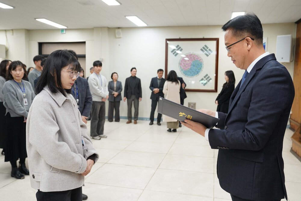 2026년 상반기 4급이하 정기인사로 정장을 입은 남성이 실내 행사장에서 한 여성에게 상장을 전달하며 내용을 읽고 있고, 여성은 안경을 쓰고 밝은 회색 카라 자켓을 입고 사원증을 착용한 채 두 손을 모은 상태로 서 있으며 뒤쪽으로 참석자들이 줄지어 서서 행사를 지켜보고 있다.