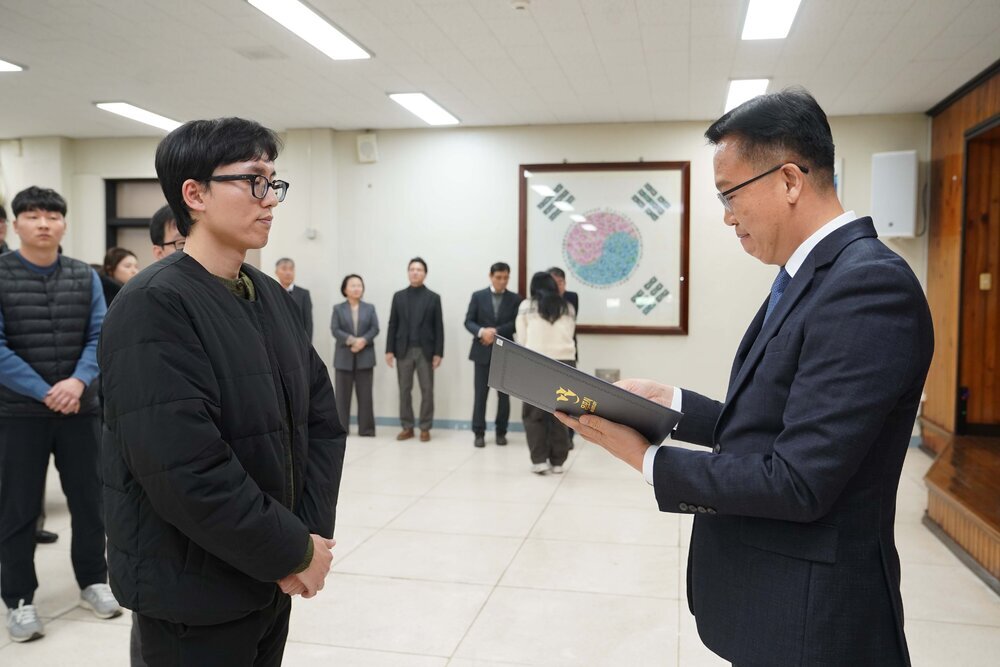 2026년 상반기 4급이하 정기인사로 정장을 입은 남성이 실내 행사장에서 한 남성에게 상장을 전달하며 내용을 읽고 있고, 남성은 검은 외투를 입고 안경을 착용한 채 두 손을 모은 상태로 서 있으며 뒤쪽에는 참석자들이 줄지어 서서 행사를 지켜보고 있다.