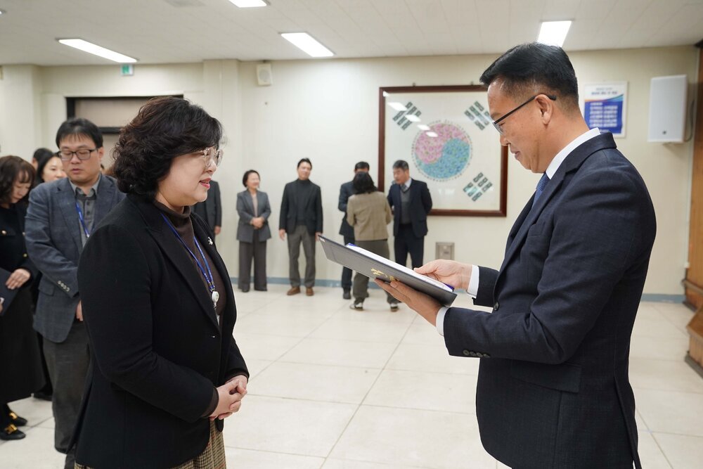2026년 상반기 4급이하 정기인사로 정장을 입고 안경을 쓴 남성이 실내 행사장에서 한 여성에게 상장을 전달하며 내용을 읽고 있고, 여성은 검은 자켓과 안경을 착용한 채 두 손을 모아 서 있으며 뒤쪽에는 여러 사람들이 줄지어 서서 행사를 지켜보고 있다.