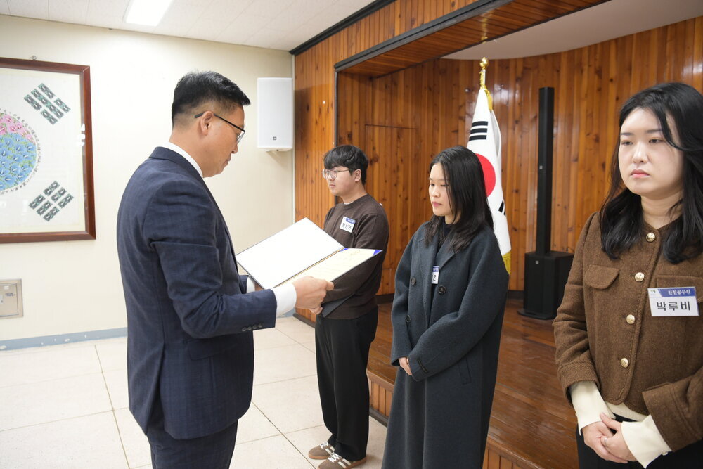 3월 정례조회에 정장을 입고 안경을 낀 남성이 검정색 롱코트를 입고 이름표가 가려진 여성에게 패를 수여하고 있다