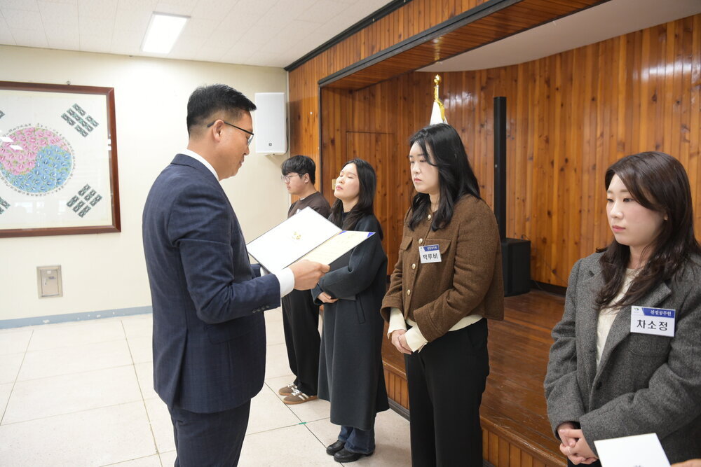 3월 정례조회에 정장을 입고 안경을 낀 남성이 갈색 짧은 코트를 입은 박루비에게 패를 수여하고 있다