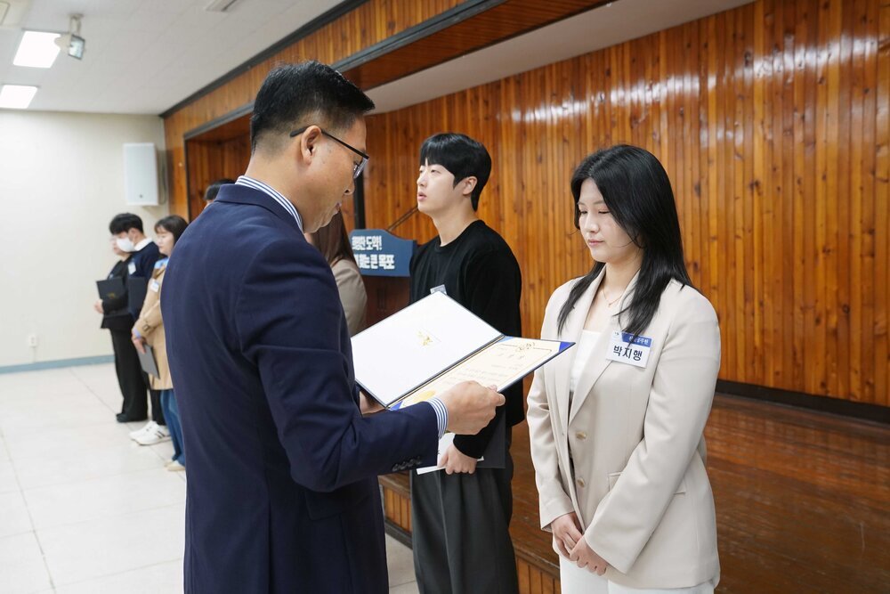 4월 정례조회에 정장을 입고 안경을 낀 남성이 밝은 베이지색 자켓을 입은 박지행에게 패를 수여하고 있다