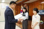 정장 차림의 관계자가 베이지색 상하의를 착용한 여성 수상자에게 상장을 전달하며 읽고 있는 모습이다.