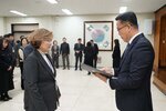 2026년 상반기 4급이하 정기인사로 정장을 입은 남성이 실내 행사장에서 한 여성에게 상장을 전달하며 내용을 읽고 있고, 여성은 밝은 갈색 파마머리에 짙은 회색 정장 자켓을 입고 사원증을 착용한 채 두 손을 내린 상태로 서 있으며 뒤쪽으로 참석자들이 줄지어 서서 행사를 지켜보고 있다.