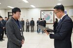 2026년 상반기 4급이하 정기인사로 정장을 입은 남성이 실내 행사장에서 한 남성에게 상장을 전달하며 내용을 읽고 있고, 남성은 안경을 쓰고 짙은 회색 보풀 니트 자켓을 입고 사원증을 착용한 채 두 손을 모은 상태로 서 있으며 뒤쪽으로 참석자들이 줄지어 서서 행사를 지켜보고 있다.