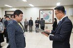 2026년 상반기 4급이하 정기인사로 정장을 입은 남성이 실내 행사장에서 한 남성에게 상장을 전달하며 내용을 읽고 있고, 남성은 안경을 쓰고 노란색 니트 위에 짙은 푸른색 자켓을 입고 사원증을 착용한 채 두 손을 내린 상태로 서 있으며 뒤쪽으로 참석자들이 줄지어 서서 행사를 지켜보고 있다.